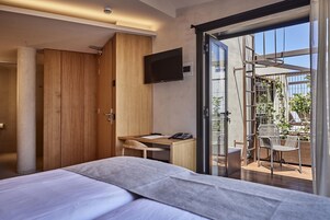 Habitación de hotel moderna con cama, escritorio y televisión. Tiene un balcón con sillas y plantas.