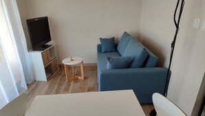 Ein Wohnzimmer mit blauem Sofa, einem kleinen runden Tisch, einem Fernseher auf einem Schrank und einem weißen Sessel.