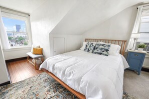 A bedroom with a bed, a nightstand, a window with a city view, a door, and a small stool.