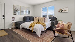 A modern bedroom with a bed, a grey sofa, a wicker chair, a small side table, and a window with a view.