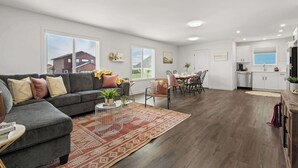 A modern living room with a gray sofa, dining area, kitchen, and large windows.