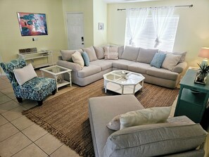 A living room with a sectional sofa, a patterned armchair, a glass coffee table, and a small side table. - Casa Azul! 11min - warm mineral springs, 25min - beaches! HEATED Pool & floats! (North Port)