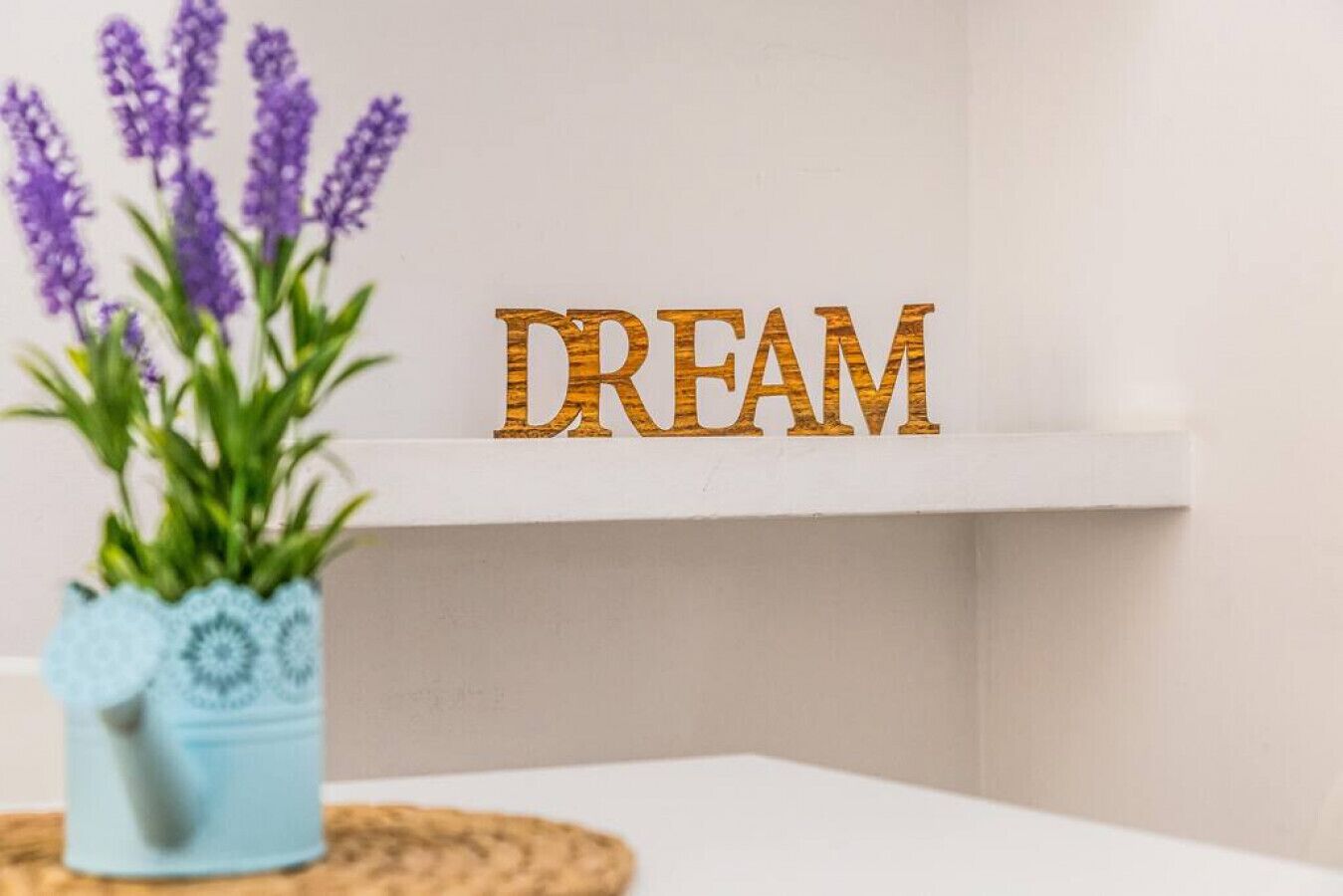 Ein weißes Regal mit der Aufschrift "DREAM" in Goldfarbe, eine blaue Vase mit Grünpflanzen und ein weißer Tisch.