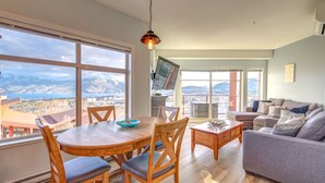 Un vaste salon doté d’une grande fenêtre offrant une vue sur les montagnes, un coin repas avec des meubles en bois et un canapé gris.