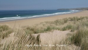 Ein Strand mit Dünen und Gras im Vordergrund, und ein Leuchtturm in der Ferne.