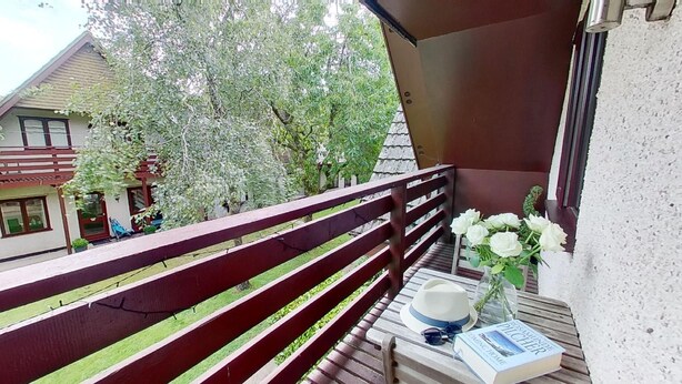 Ein Balkon mit Holzgeländer, einem Tisch mit einem Buch, einem Hut, Sonnenbrillen und einer Vase mit weißen Blumen.
