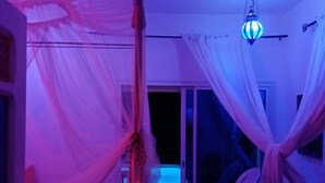 A room with a canopy bed, a chair, a table, and a view of the outside. - Dar Soulayma vacation home (Fatou houmet souk Djerba)