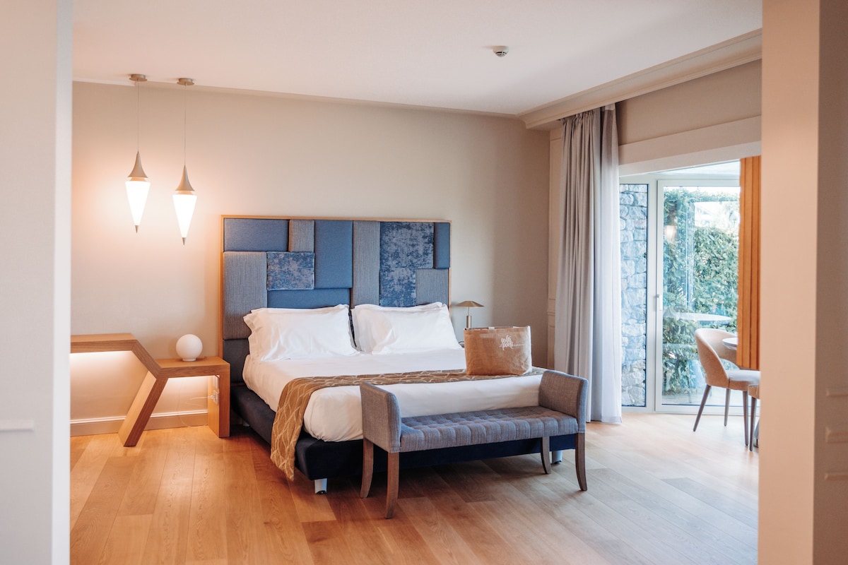 A modern bedroom with a large bed, bedside tables, a bench, and a view of greenery through a glass door.