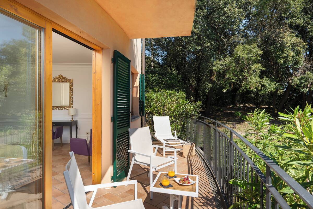 Un balcon avec des chaises blanches, une table avec des fruits et du jus, et une vue sur les arbres.