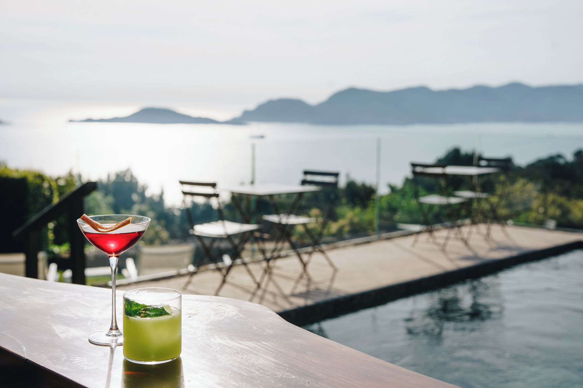 Angolo bar con vista sulla piscina, con un bicchiere da martini e un cocktail verde, e posti a sedere all'aperto con tavoli e sedie.