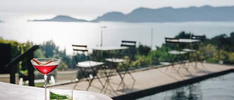 Zona de bar con vistas a la piscina, incluyendo una copa de martini y un cóctel verde, y mobiliario exterior con mesas y sillas.