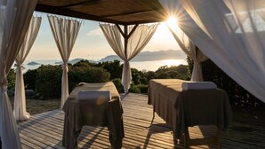 Una terraza de madera con dos camillas de masaje, cada una con una toalla, bajo una pérgola con cortinas translúcidas.