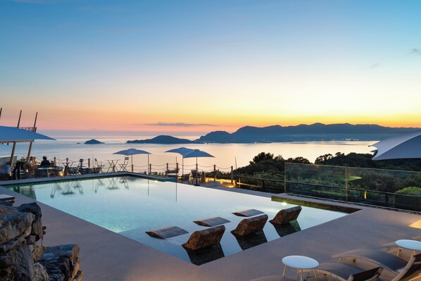 An infinity pool with lounge chairs and umbrellas, overlooking a body of water and distant mountains.
