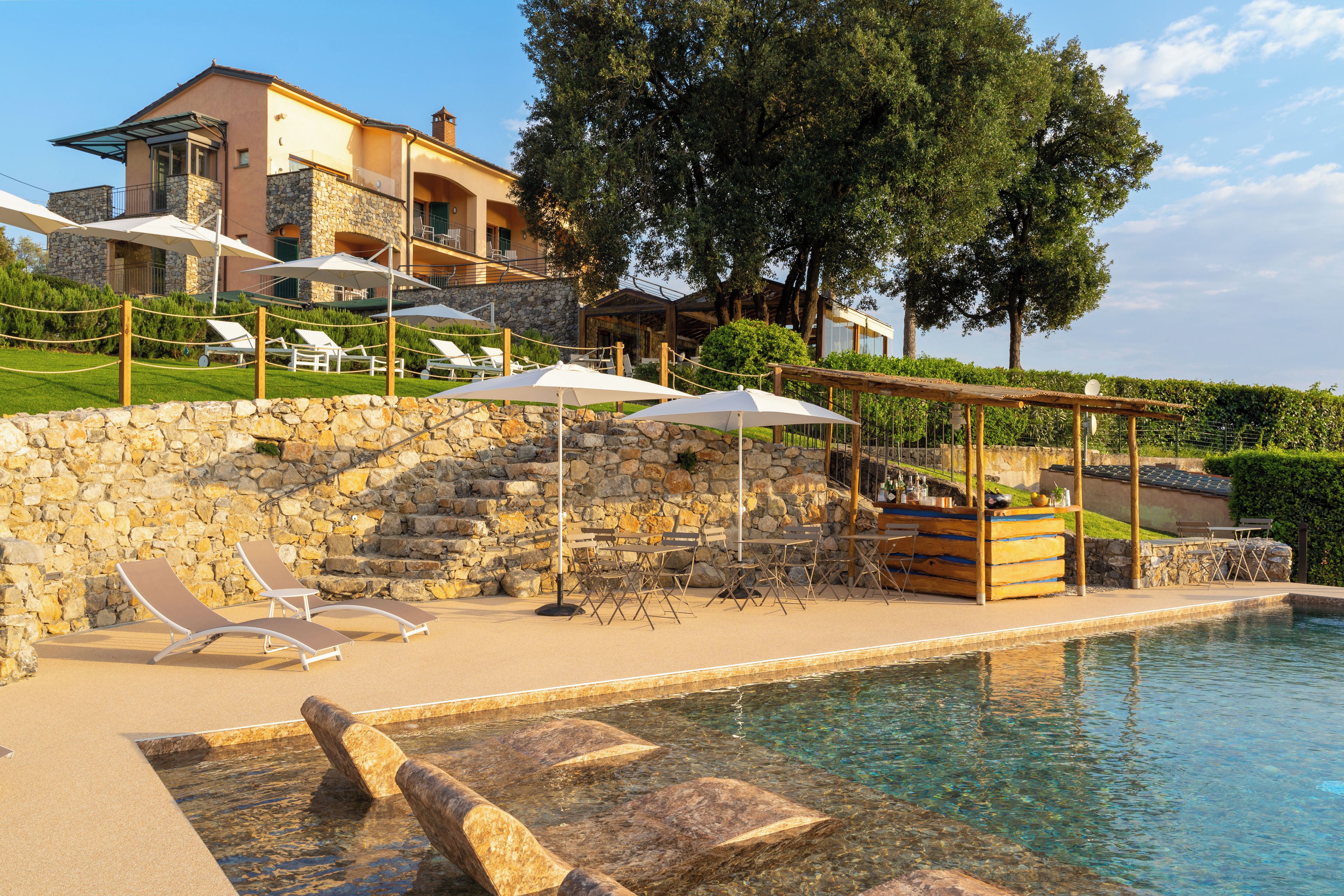 Un espace piscine avec des chaises longues et des parasols, un mur en pierre et un bâtiment avec un balcon en arrière-plan.