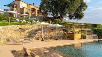 Un espace piscine avec des chaises longues et des parasols, un mur en pierre et un bâtiment avec un balcon en arrière-plan.