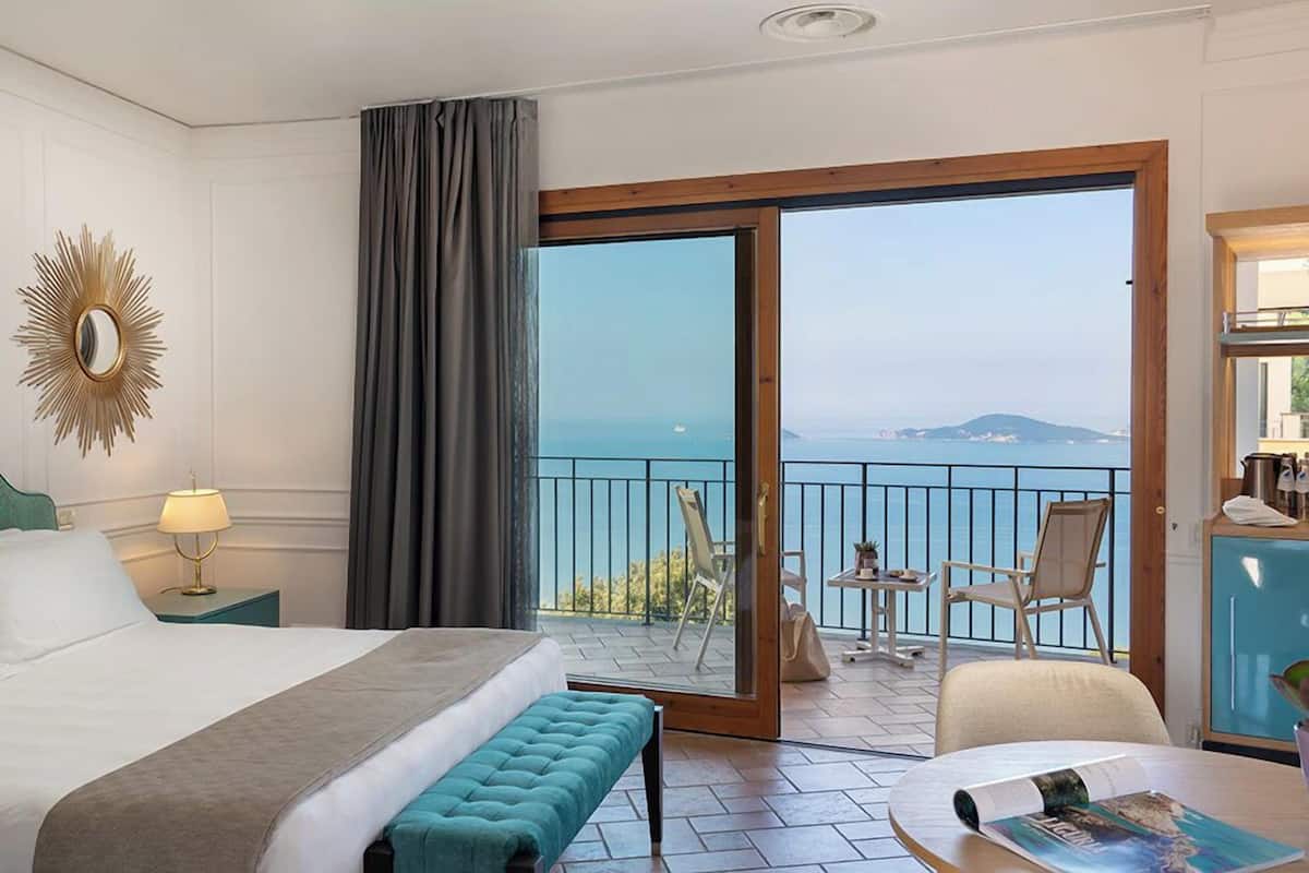 Une chambre d’hôtel avec un lit, une banquette, une table avec un magazine, une lampe, un miroir et un balcon avec des chaises et une vue sur la mer.
