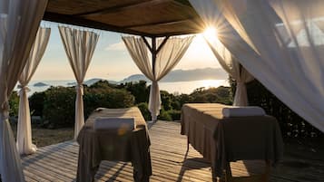Una terraza de madera con dos camillas de masaje, cada una con una toalla, bajo una pérgola con cortinas translúcidas.