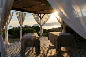 En træterrasse med to massagestole, hver med et håndklæde, under et halvtag med lette gardiner.