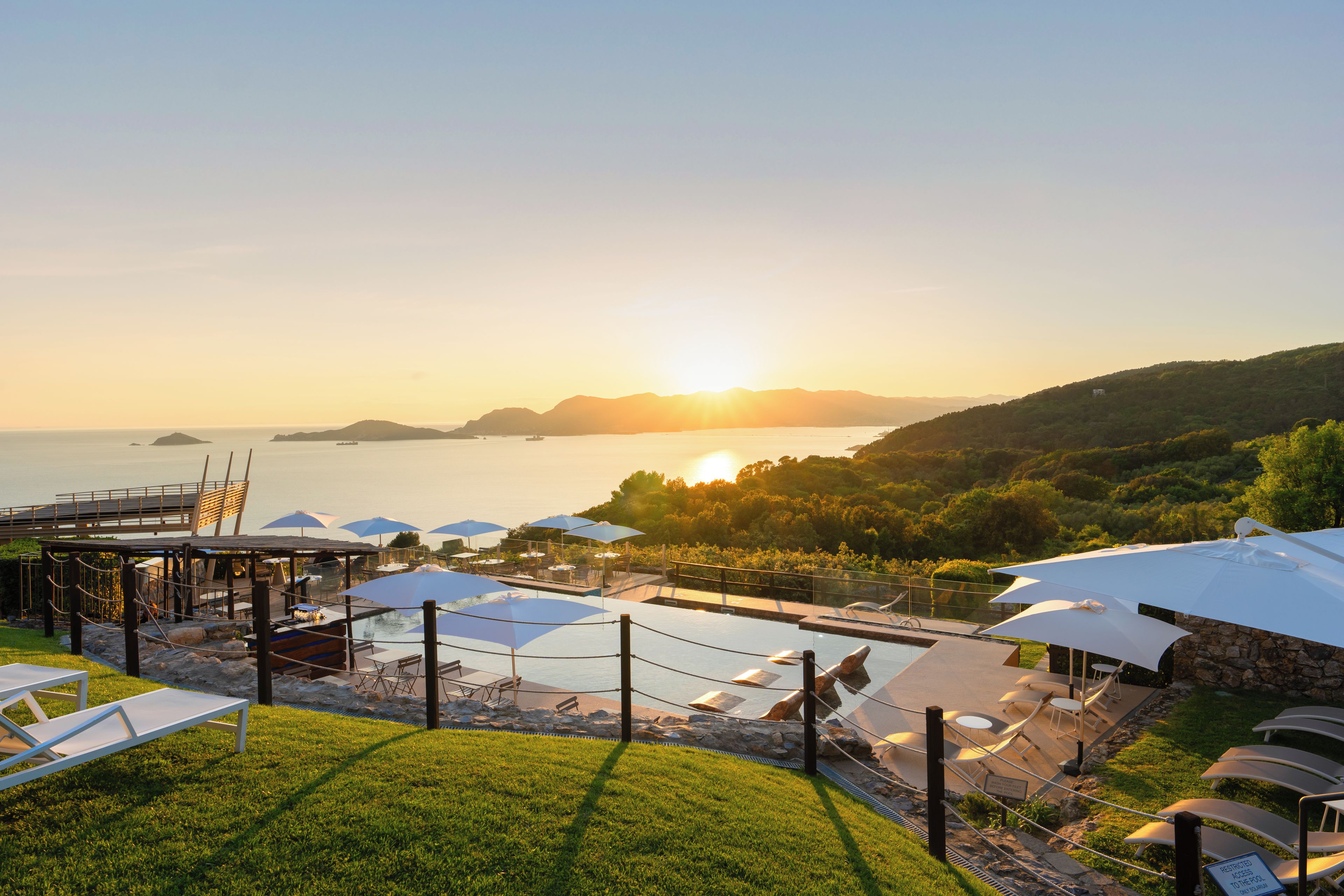 A resort pool area with lounge chairs and umbrellas, overlooking a body of water and hills during sunset.