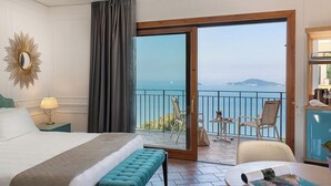 Habitación de hotel con cama, reposabajares, una mesa con una revista, lámpara, espejo y balcón con sillas y vista al mar.