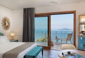A hotel room with a bed, a bench, a table with a magazine, a lamp, a mirror, and a balcony with chairs and a view of the sea.