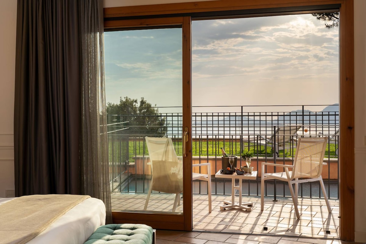 A hotel room with a large sliding glass door leading to an outdoor terrace with a view of the sea and mountains.