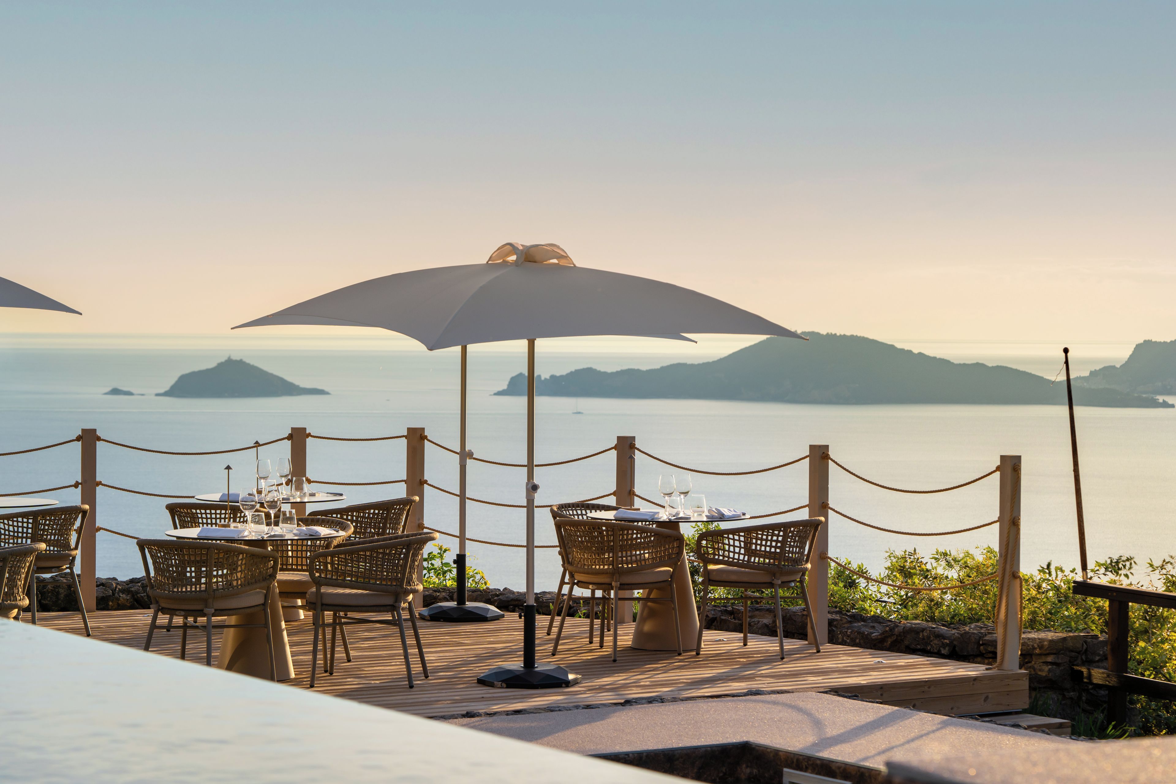 Un espace de restauration en bord de mer, avec des tables et des chaises, une barrière faite de cordes, et une vue sur l’océan et les îles lointaines.