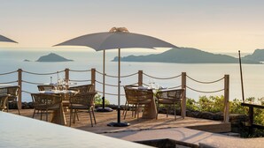 Un área de comedor junto al mar con mesas y sillas, una barrera de cuerda y vistas al océano y a islas distantes.