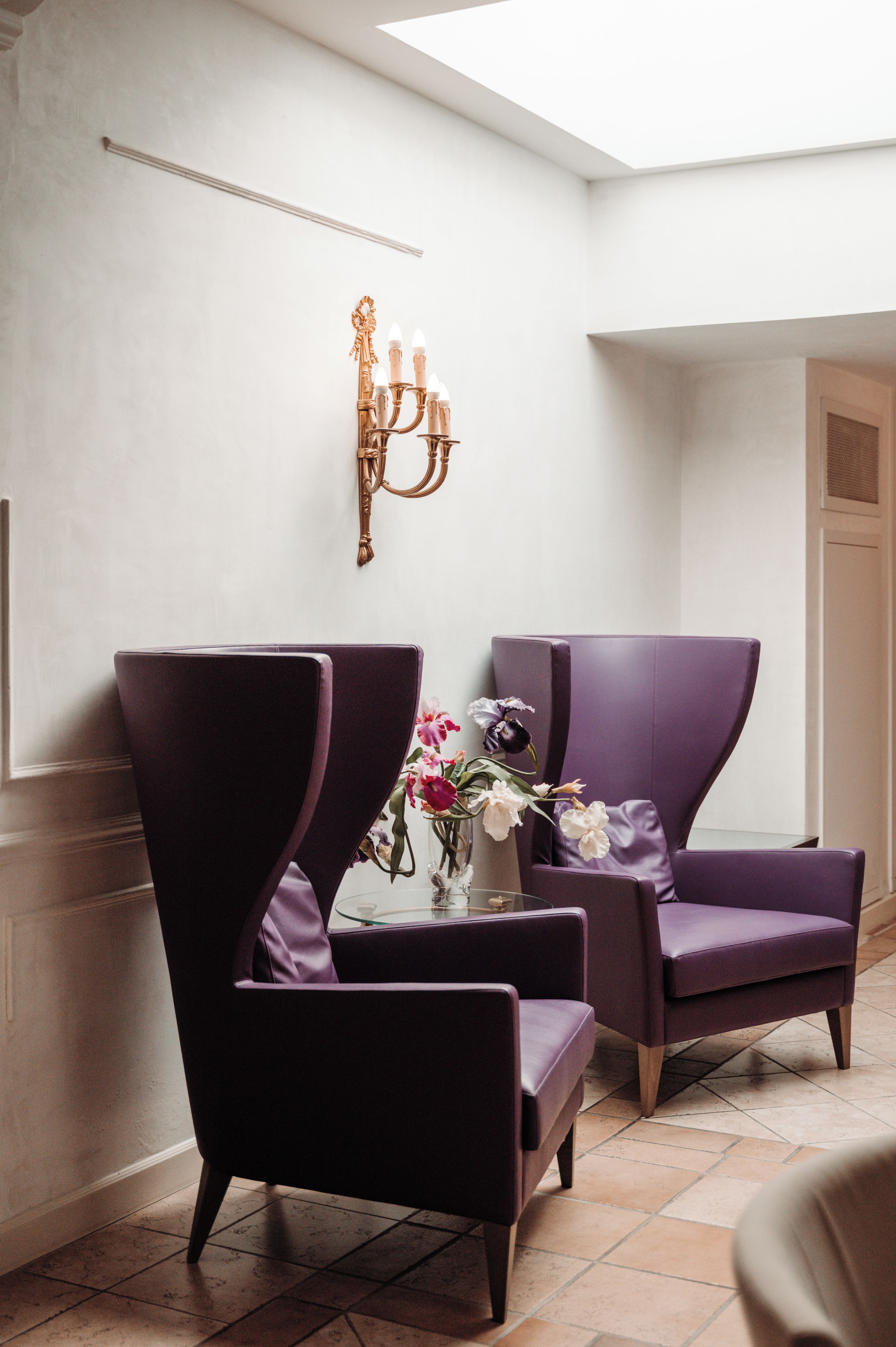 A room with two purple armchairs, a small table with a vase of flowers, and a wall-mounted candelabra.