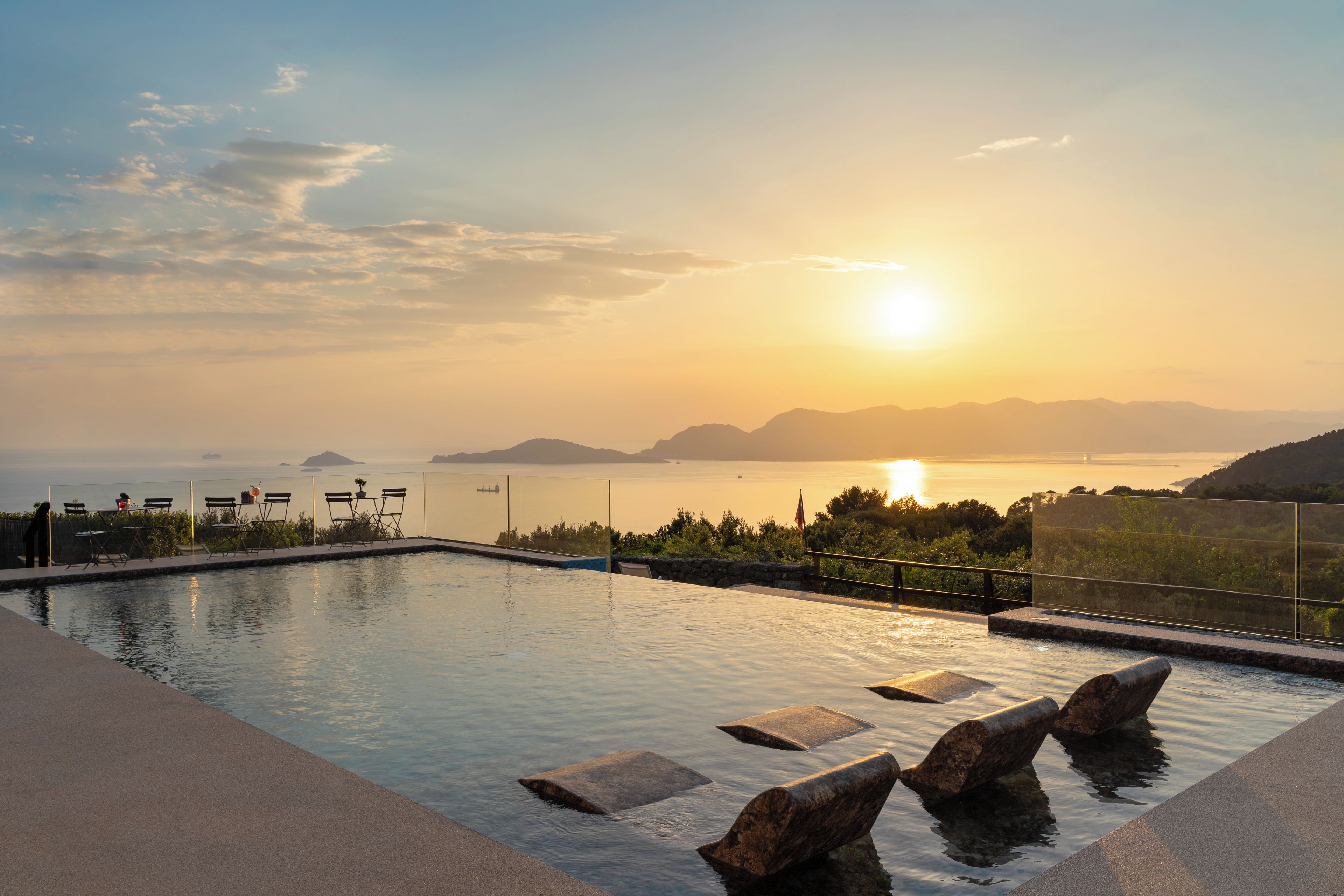 A swimming pool with a view of the sunset over the water, surrounded by chairs and a railing.