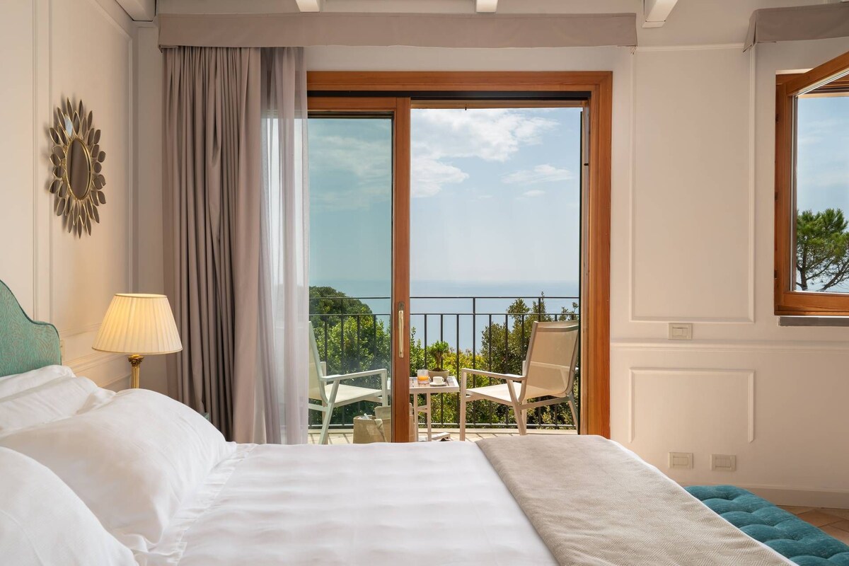 A bedroom with a bed, a lamp, a view of the sea, and a balcony with a table and chairs.