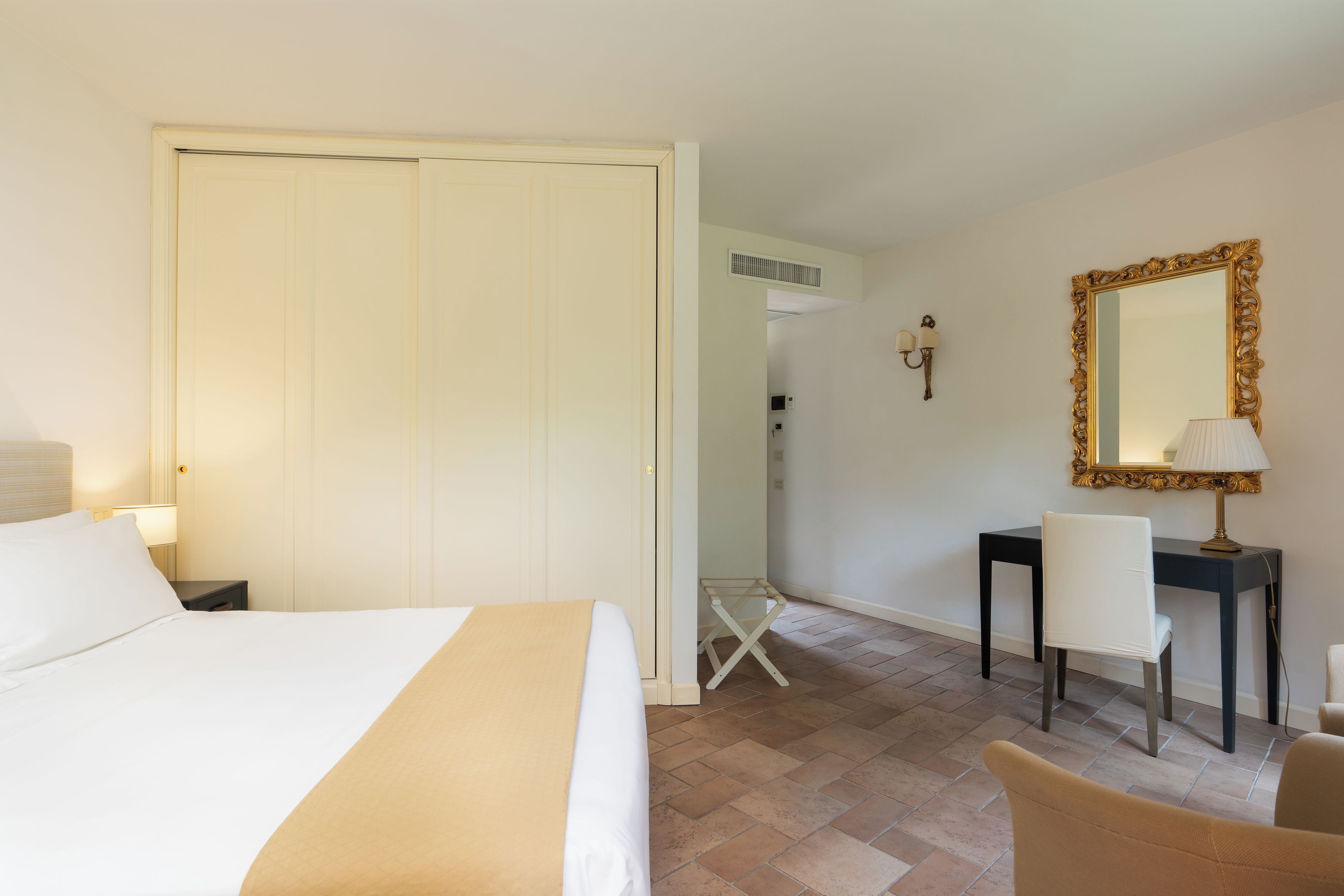 A hotel room with a bed, a desk, a chair, a mirror, and a lamp.