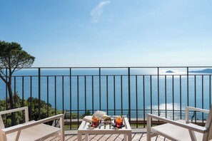 Un balcón con vistas despejadas al mar, dos sillas, una mesita con bebidas y un libro, y una barandilla metálica.