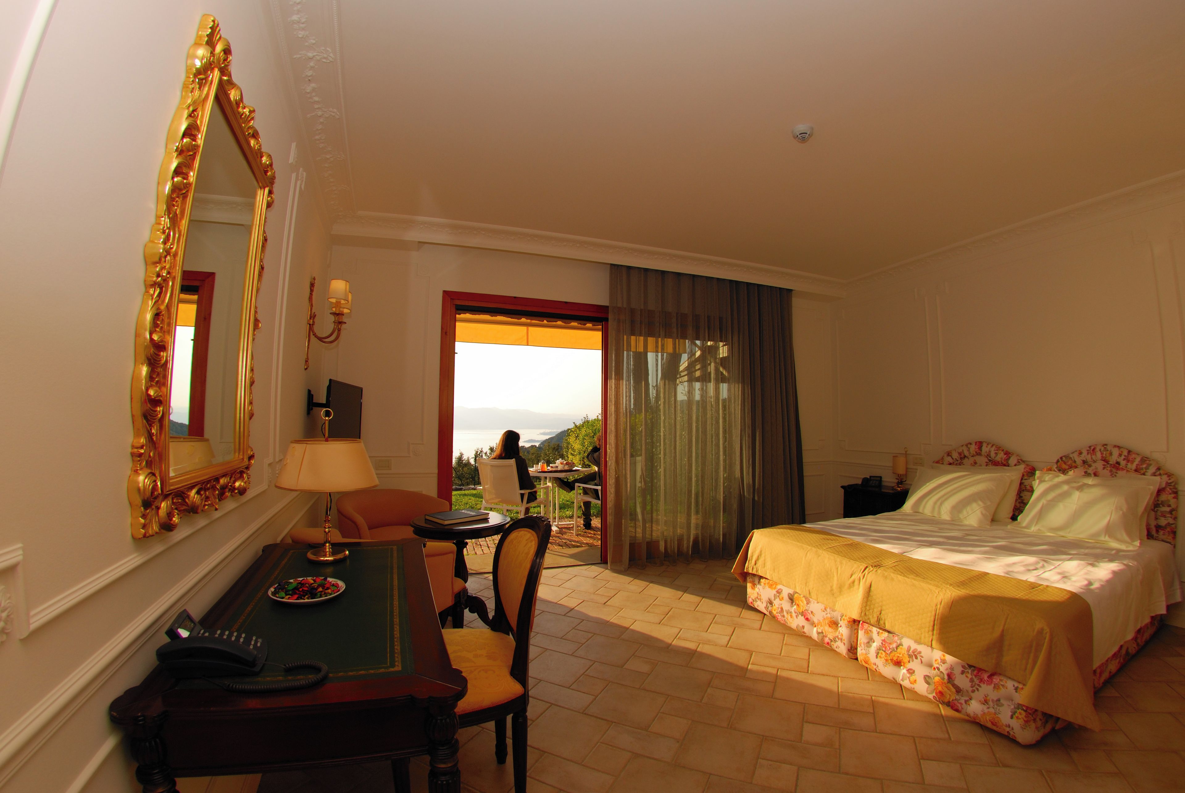 A hotel room with a bed, a desk with a telephone, a chair, a table with a lamp, and a view of the outdoors.