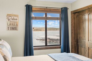 A bedroom with a large window overlooking a dock and water, a bed with a blue blanket, and a wooden door.