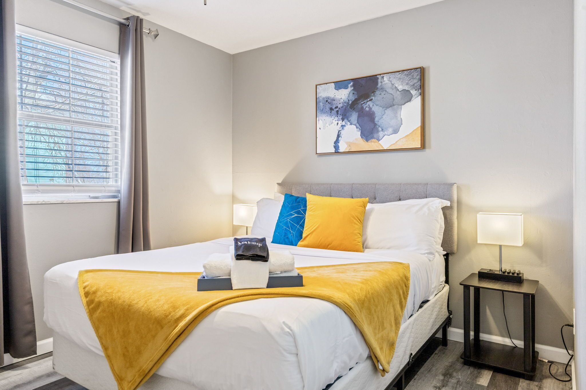 A hotel room with a bed, a nightstand, a lamp, a painting on the wall, and a window with curtains.