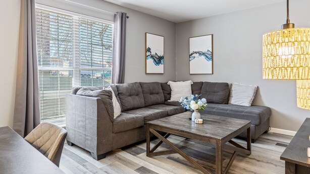 A modern living room with a sectional sofa, a wooden coffee table, and a large window with blinds.