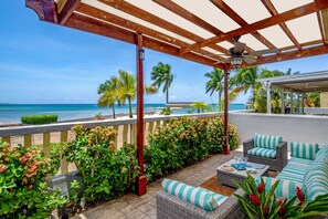 Un espace extérieur aménagé, abrité et doté de mobilier en osier et de coussins à rayures, offrant une vue sur une plage bordée de palmiers et sur l’océan.