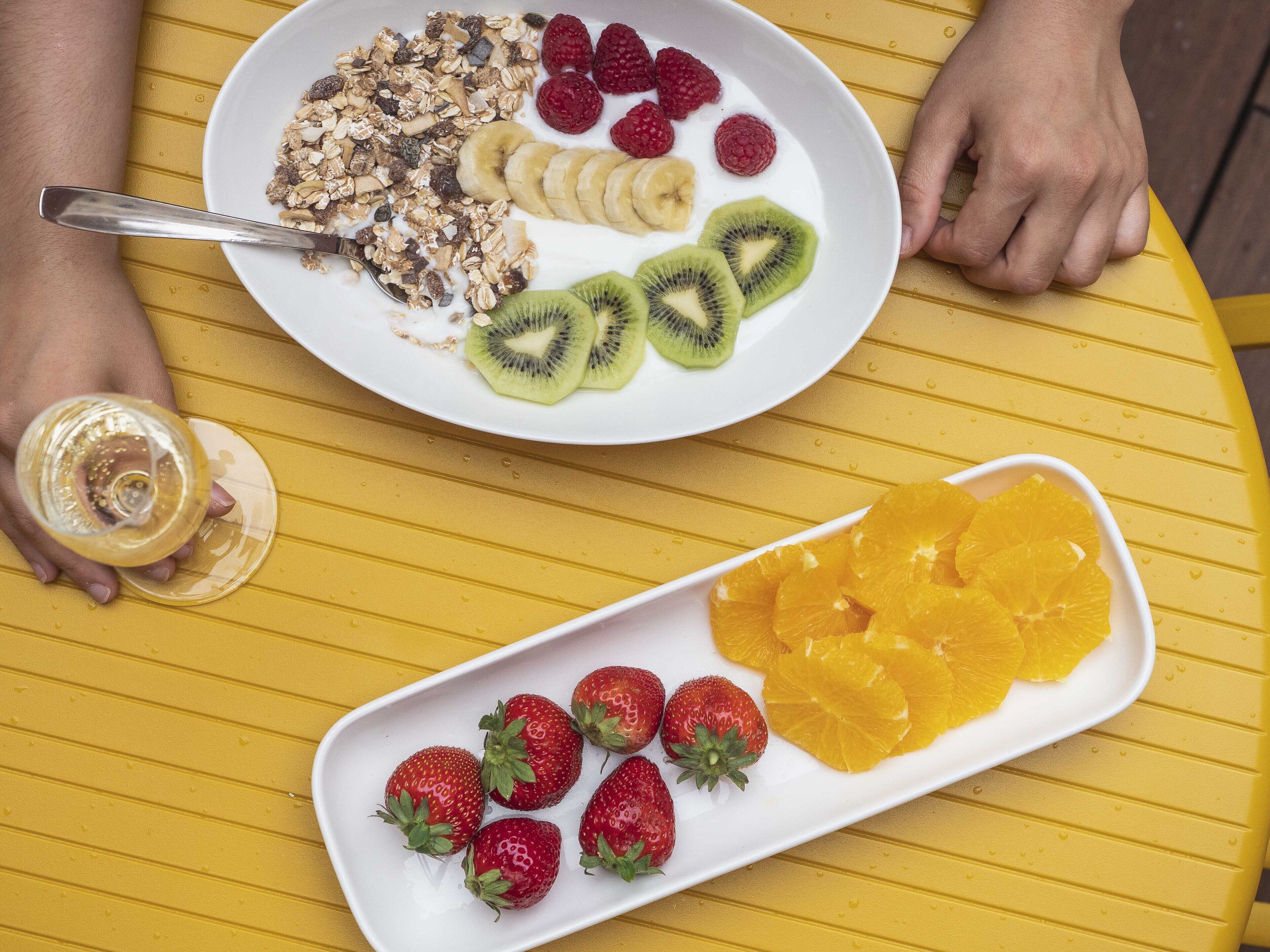 A breakfast setting with a plate of cereal, fruits, and a glass of juice on a yellow table.