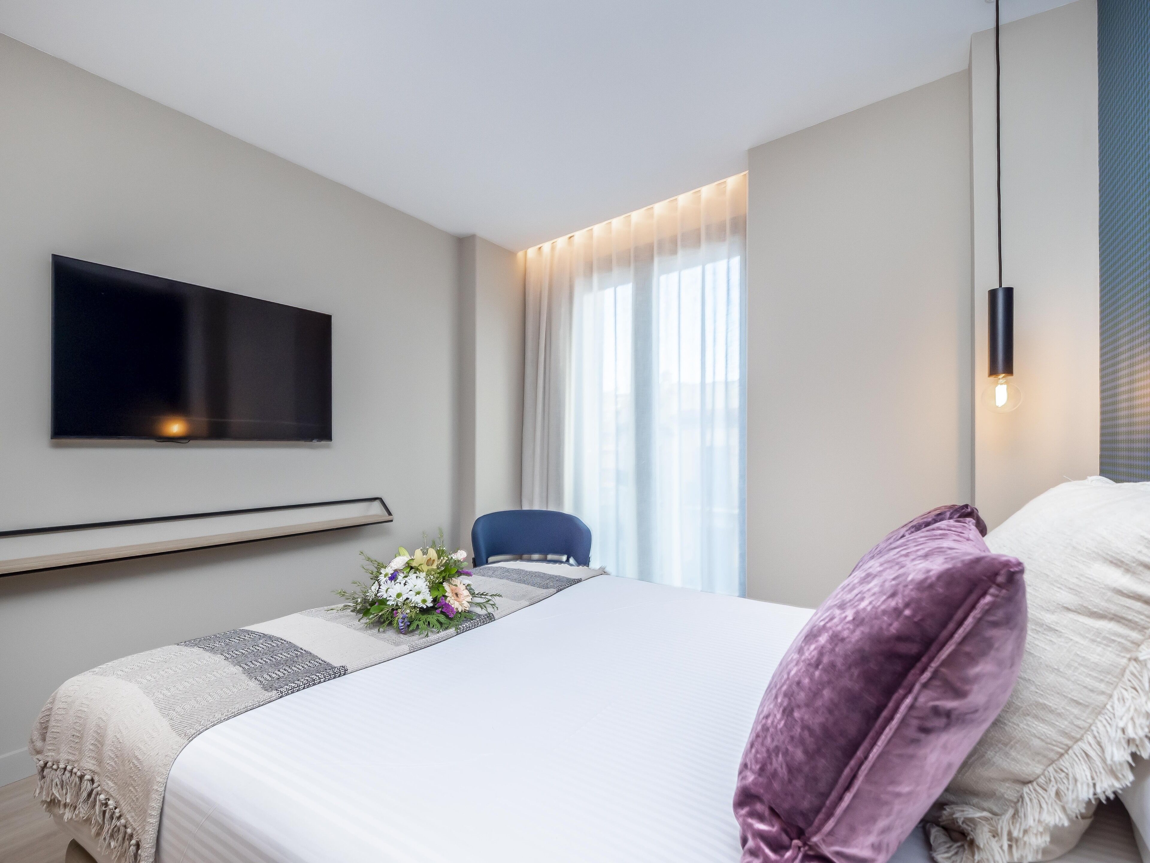 Una habitación de hotel moderna con una cama grande, un televisor de pantalla plana montado en la pared, una mesita con un centro de mesa floral y una ventana con cortinas.