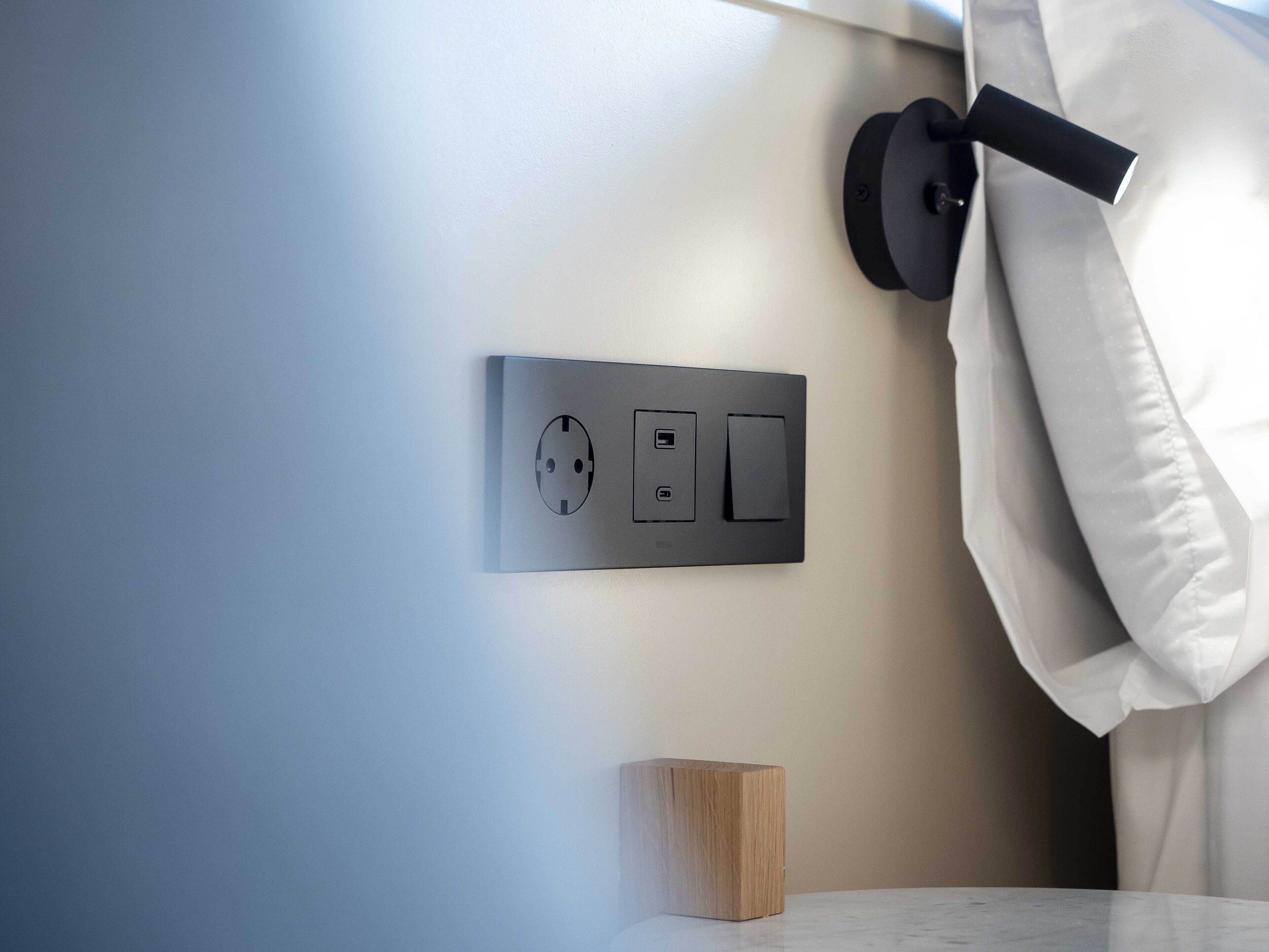 A modern wall-mounted electrical outlet with a USB port and a switch, a wooden block, and a white curtain.