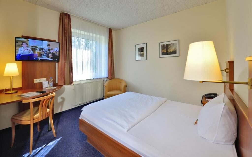 A hotel room with a bed, a desk with a chair, a television, and two framed pictures on the wall.