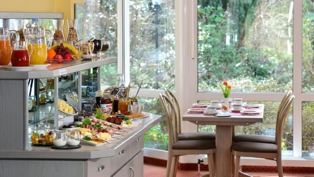Ein reichhaltiges Frühstücksbuffet mit Speisen und Getränken, ein Esstisch mit Stühlen und Blick auf Grünflächen durch große Fenster.