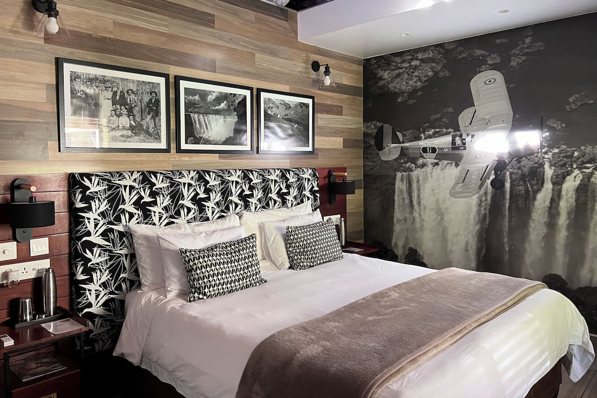 A modern hotel room with a wooden headboard, a large bed with patterned pillows, a nightstand with a lamp, and a wall-mounted light fixture.