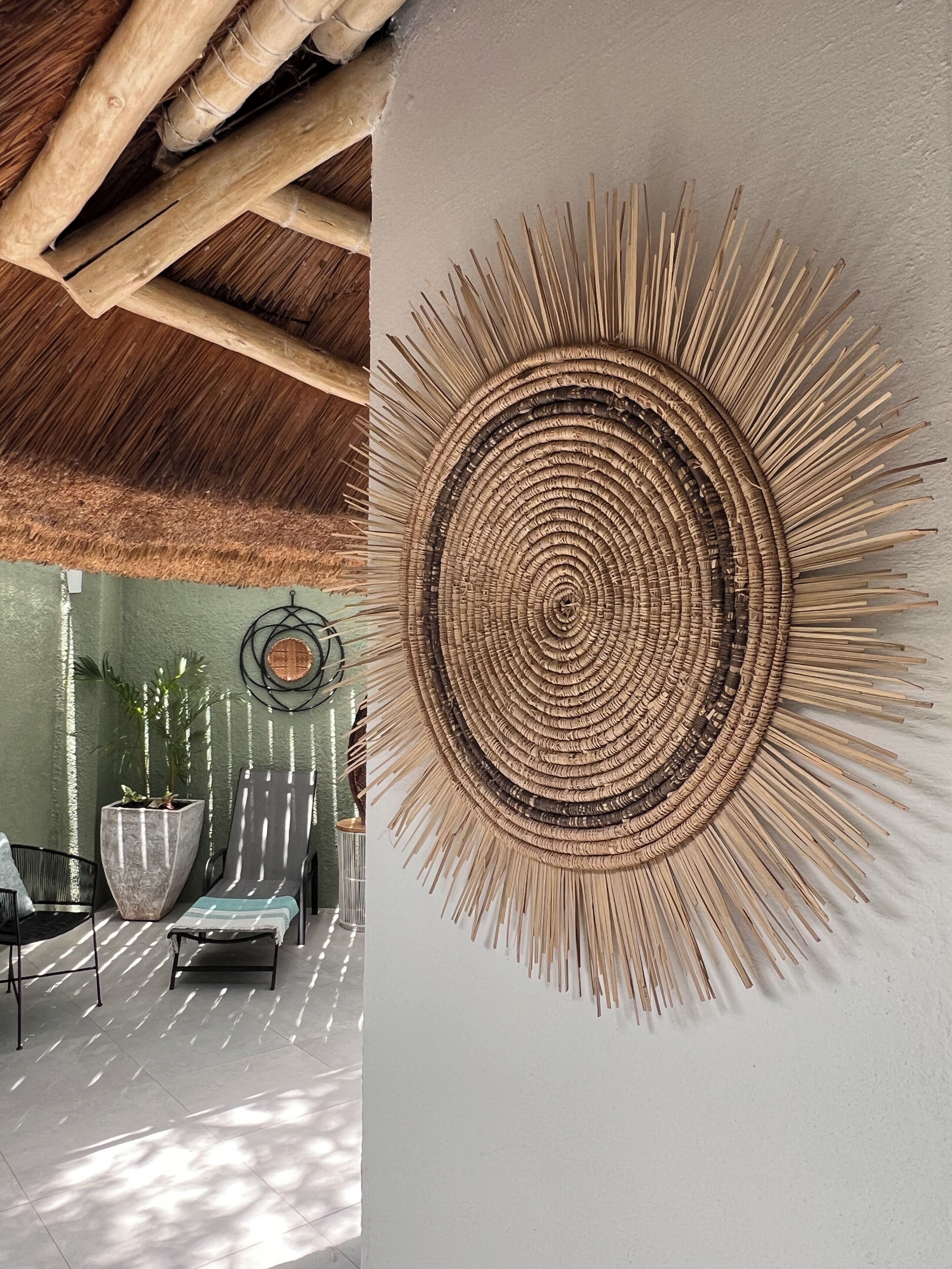 A room with a thatched ceiling, a large woven wall art piece, and a seating area with a potted plant and a metal chair.