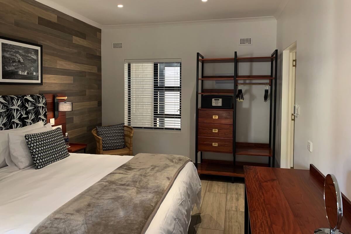 A bedroom with a bed, a wooden wardrobe, a chair, a desk, and a window with blinds.