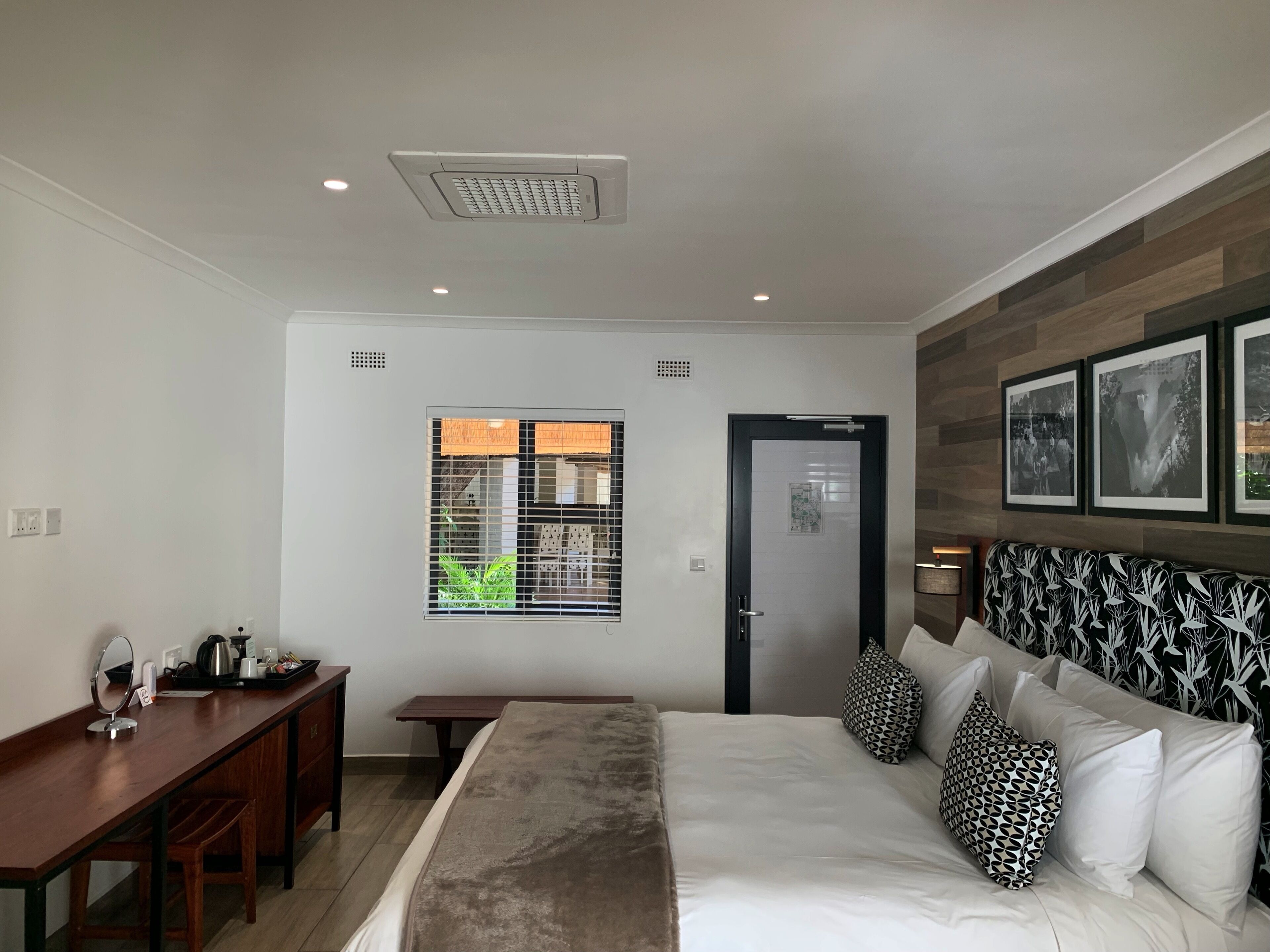 Une chambre d’hôtel moderne avec un grand lit, un bureau, une fenêtre avec des stores, une porte et des tableaux encadrés au mur.