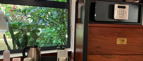 A hotel room with a wooden desk, a tray with toiletries, a window with a view of greenery, and a minibar with a minibar key.
