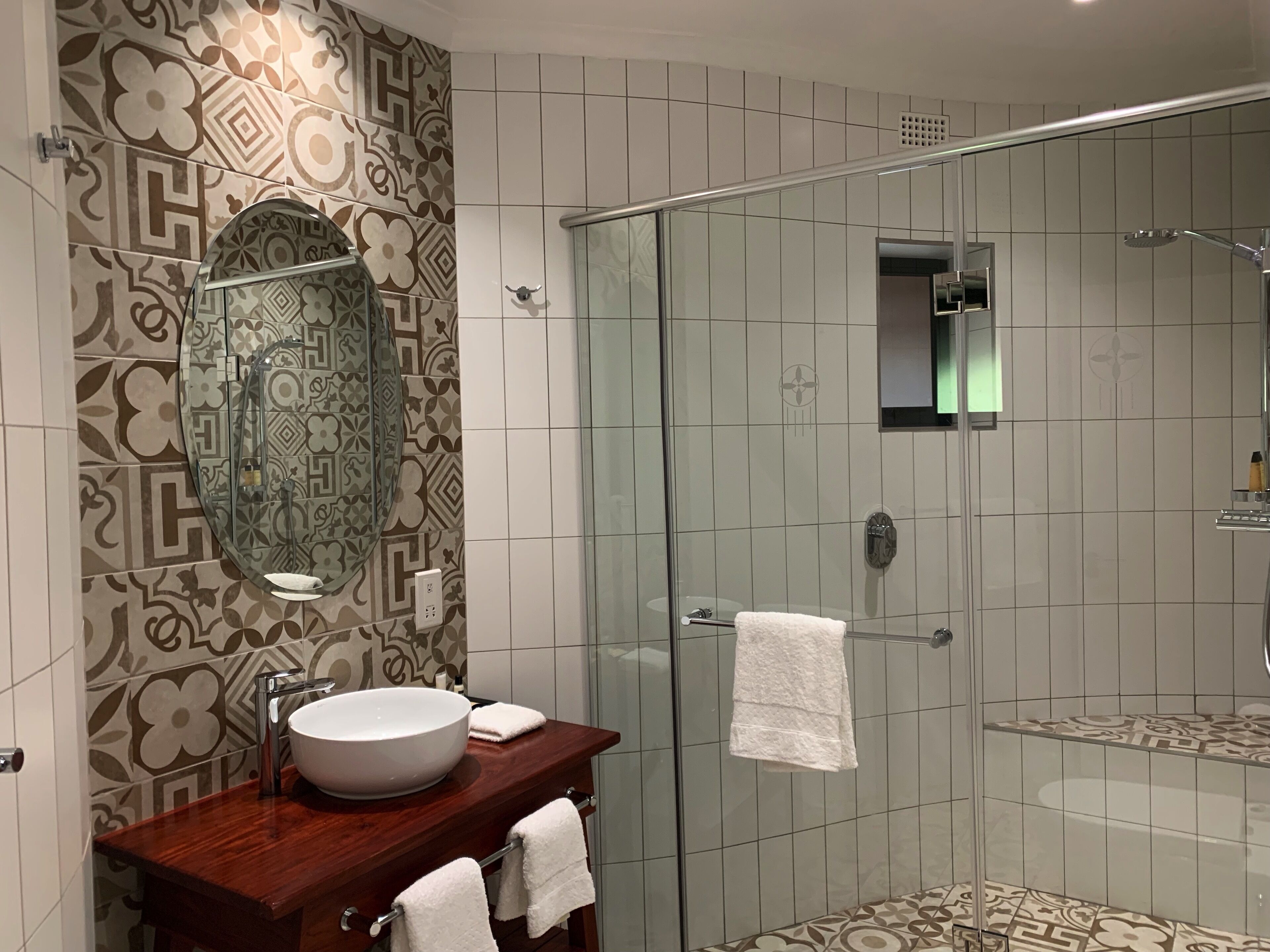 A bathroom with a large mirror, a wooden vanity, a glass shower enclosure, and patterned tiles.