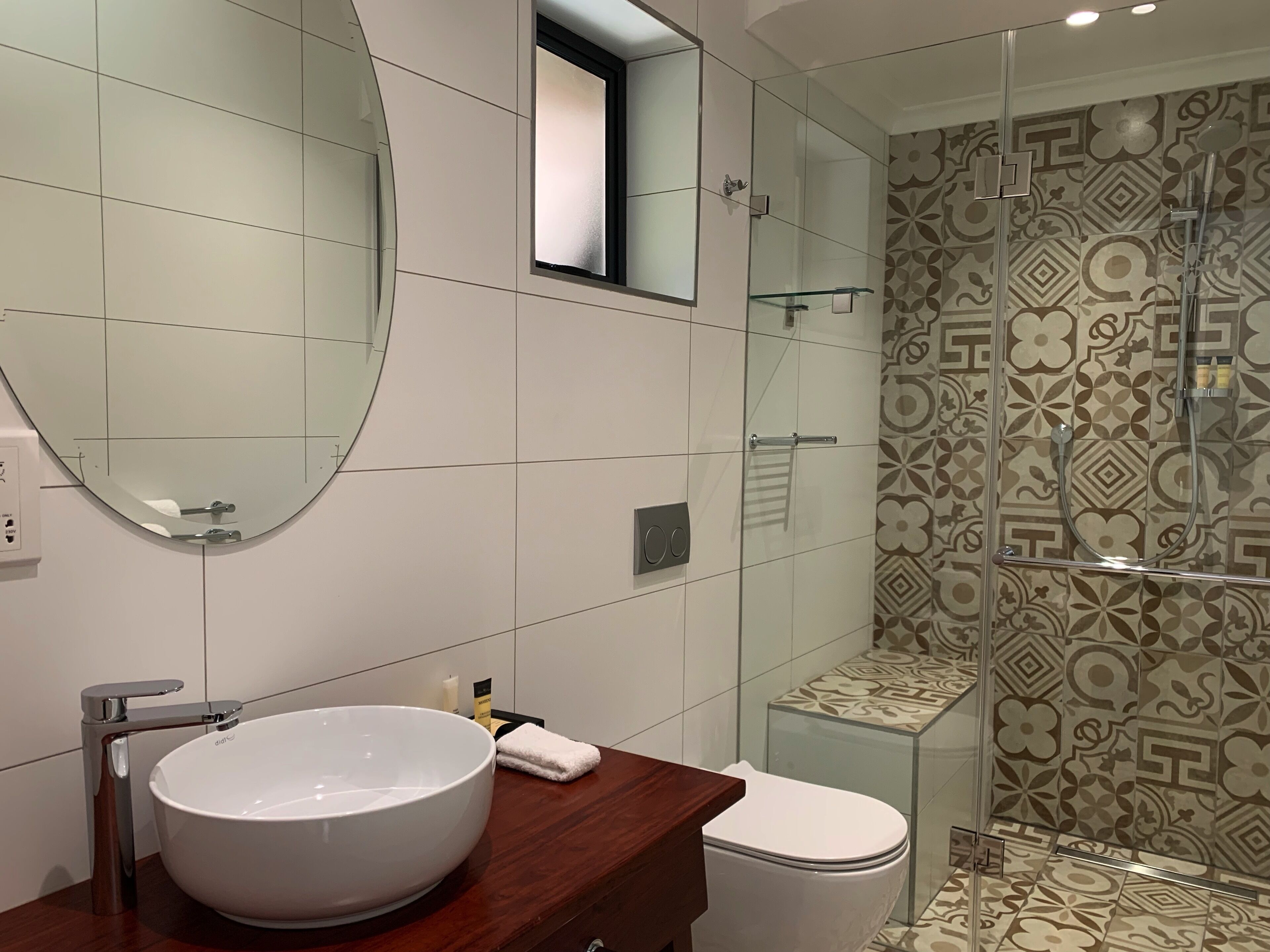 Een moderne badkamer met een grote, ovale spiegel, een witte wastafel, een douchecabine met glazen wanden en betegeling met een patroon.